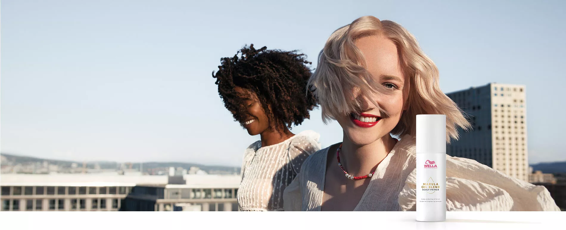 Girl with short blonde hair and girl with textured hair photographed on a rooftop, and bottle of Marula Oil Blend Scalp Primer in the foreground