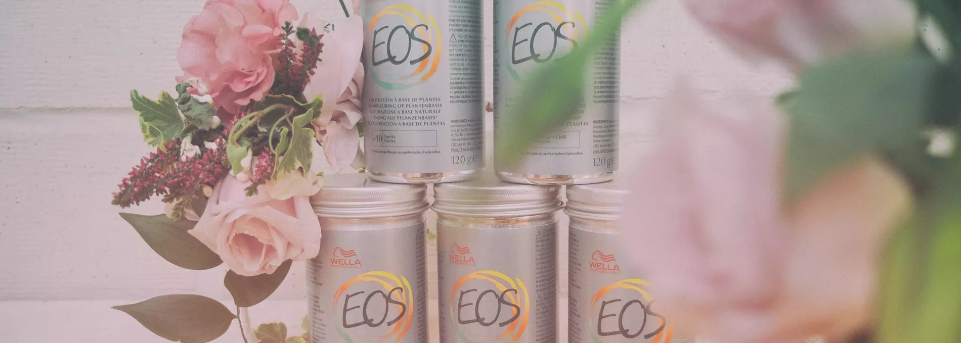 Five silver EOS plant based hair dye cans stacked up on top of each other, next to pink flowers.