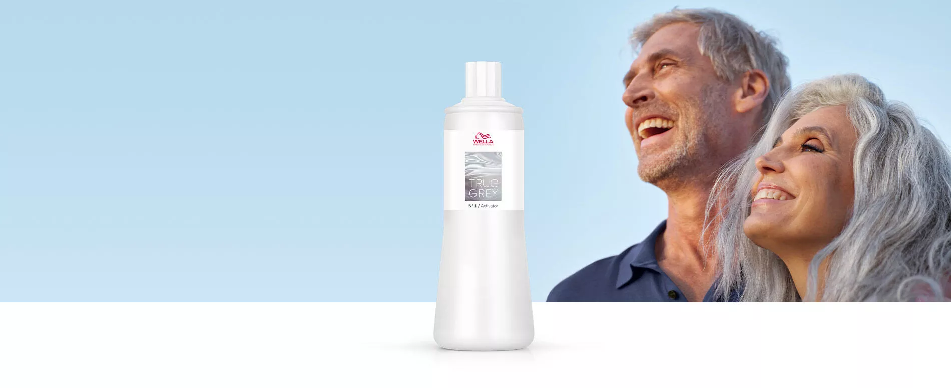 Un hombre y una mujer con cabello gris natural riéndose frente al sol con una botella de activador True Grey al fondo